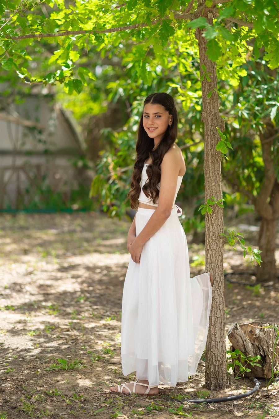 Young girl in a white dress smiling under green trees.